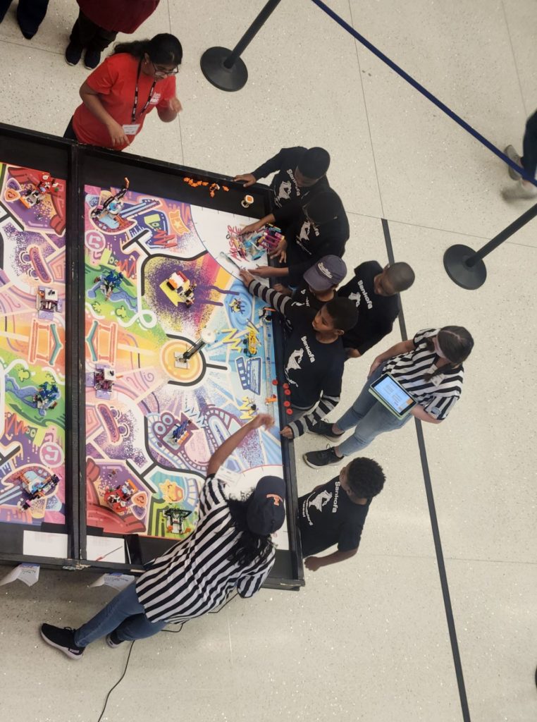 Overhead view of the team gathered around a large game table. There are referees in striped shirts looking on.