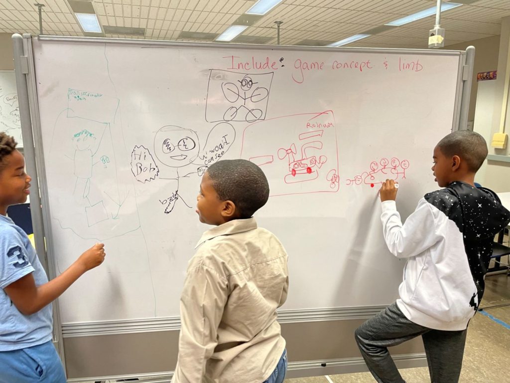 Three Black boys are standing at a large white board, drawing plans for a robot.