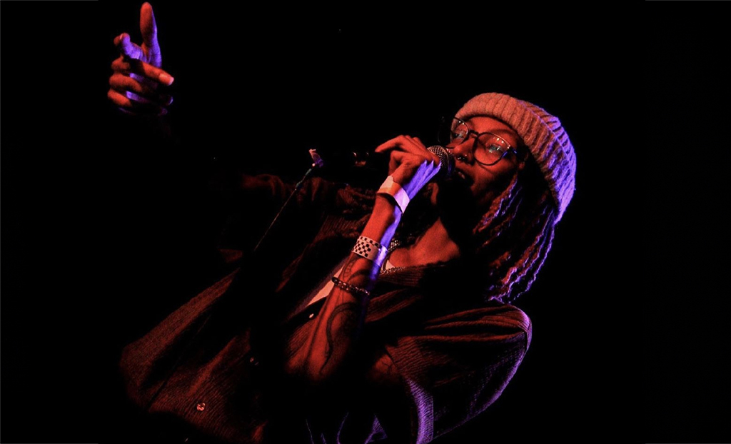 The image depicts a performer on stage under red stage lighting. The artist is wearing glasses, a beige knitted beanie, and a dark long-sleeved shirt. They are holding a microphone to their mouth with one hand and extending their other hand towards the audience, with their fingers pointing out as if making a connection with someone in the crowd. The overall atmosphere is moody and focused, with the performer illuminated against a dark background, enhancing the sense of live performance engagement.