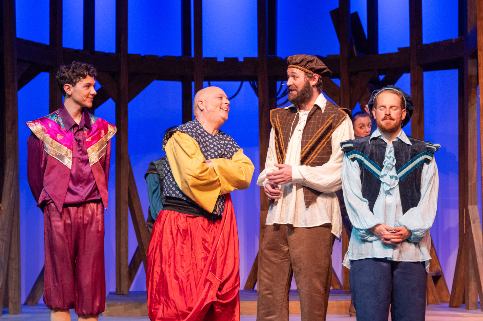 Four actors in Rosencrantz & Guildenstern are dead. They are four white men; the two on the left are looking toward the third man from the left, who looks back at them. The fourth man, on the right of the image, faces forward with his eyes closed. They are all wearing vaguely Shakespearean dress. There is a wooden structure behind them; the stage is lit blue. Actors are: Owen Henderson (The Player), David Dillman (Alfred), Anthony DeGregorio (Guldenstern), Douglas Malcolm (Rosencrantz)