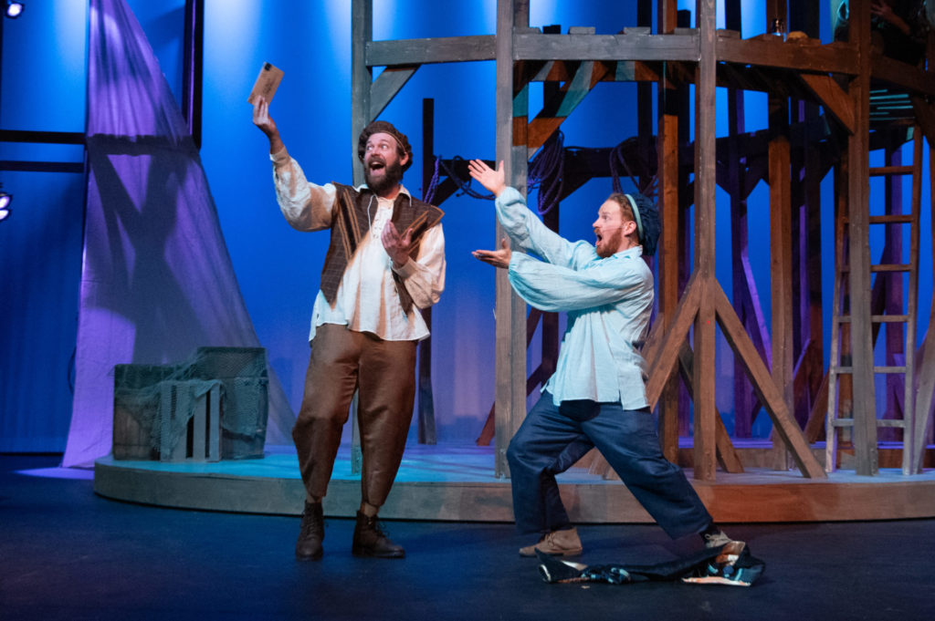 Two white men on stage for Rosencrantz and Guildenstern Are Dead. On the left, a man wearing brown trousers and a brown vest, and a white shirt. On the right, a man wearing dark blue pants and a light blue shirt. The man on the left holds a letter up, and the man on the right gesticulates toward it.