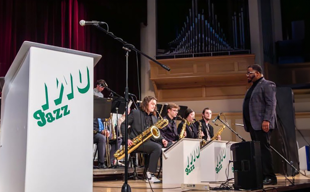 A jazz ensemble is seated on a stage with a large pipe organ in the background. In the foreground on the left, a podium with the word "Jazz" prominently displayed is positioned next to a microphone stand. A man stands to the right of the podium, facing the ensemble. He is dressed in a dark suit and appears to be directing or preparing to address the group. The band members are seated with their instruments, including saxophones and a trombone, focused on sheet music stands in front of them. The setting suggests a formal concert or performance in a venue designed for musical events.