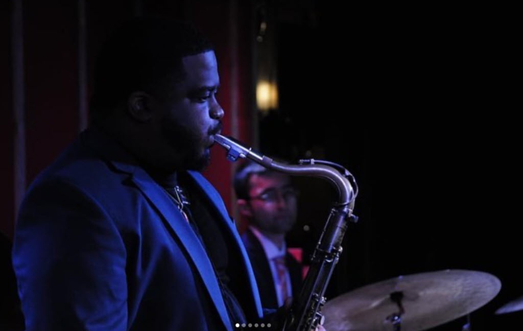 A man is seen in the foreground playing a saxophone, his focus intent upon the instrument. He is wearing a dark blazer over a formal ensemble, suggesting a jazz or formal music event setting. In the background, another person, slightly out of focus, appears to be seated at a drum set, hinting at a band or ensemble performance. The lighting is subdued with a spotlight effect, which typically indicates a stage or a performance venue environment.