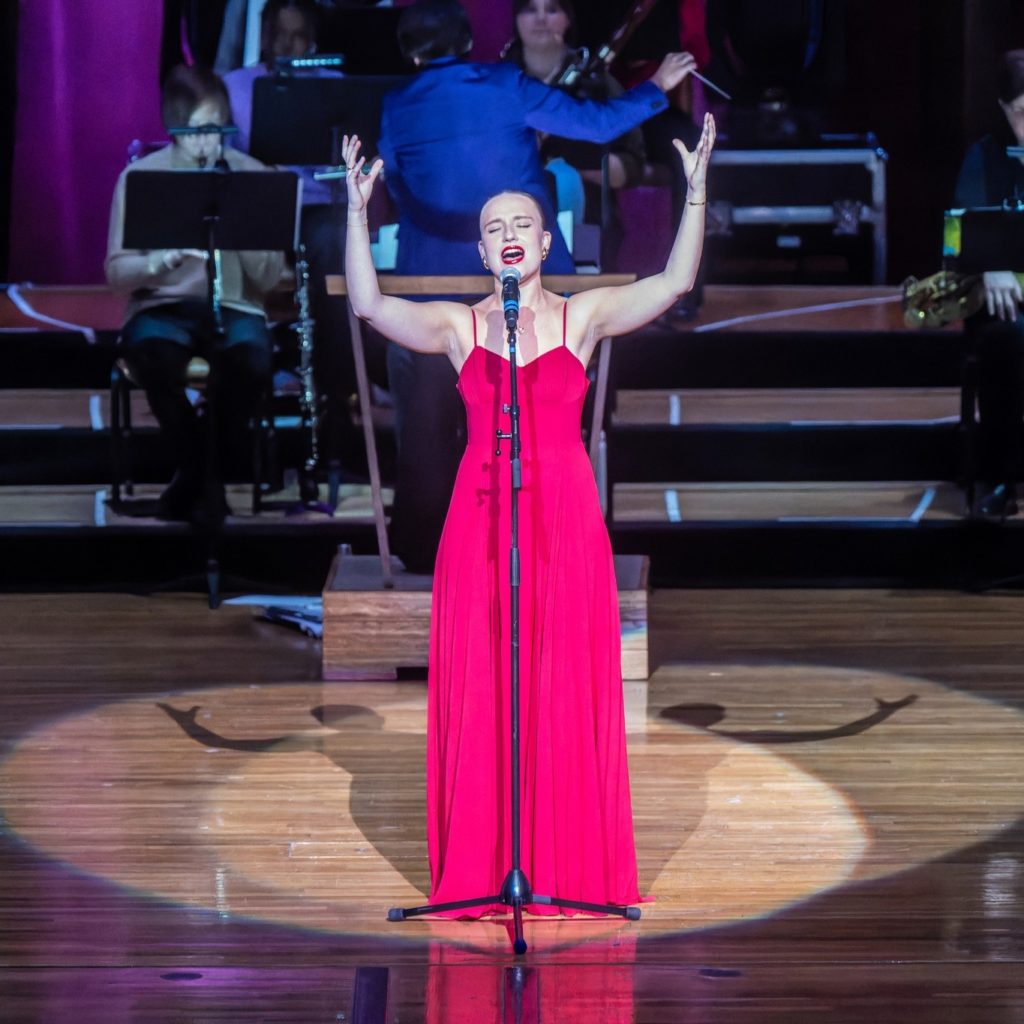 A white woman wearing a long pink gown with spaghetti straps sings at a microphone stand on a stage. Her arms are raised and her eyes are closed. The spotlight is on her.