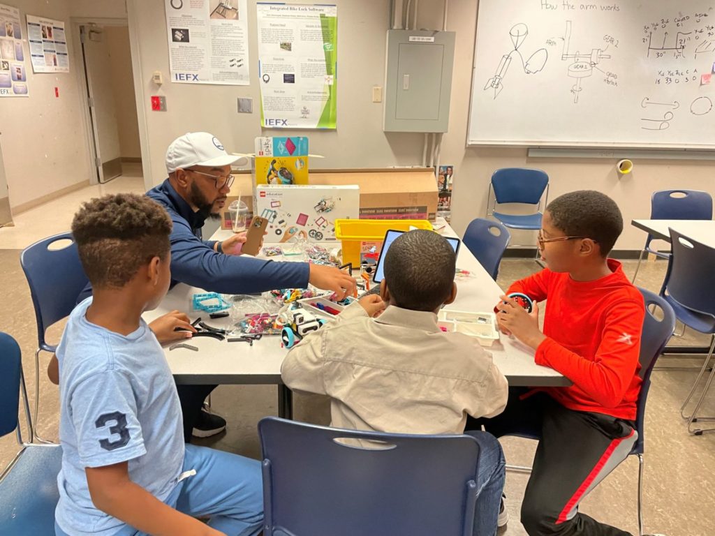 A Black man in a white cap, and three young Black boys are seated around a table, looking at a Lego kit.