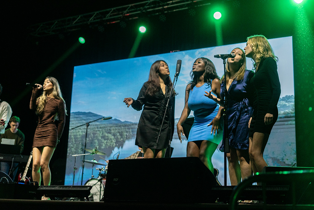 Five women stand on stage performing at a concert. They are dressed in evening attire, with one in a brown long-sleeve mini dress, another in a black dress, the third in a blue sleeveless dress, and the fourth in a black sleeveless dress. Behind them is a screen displaying a serene landscape, and various musical instruments, including keyboards and a drum set, are partially visible on stage.