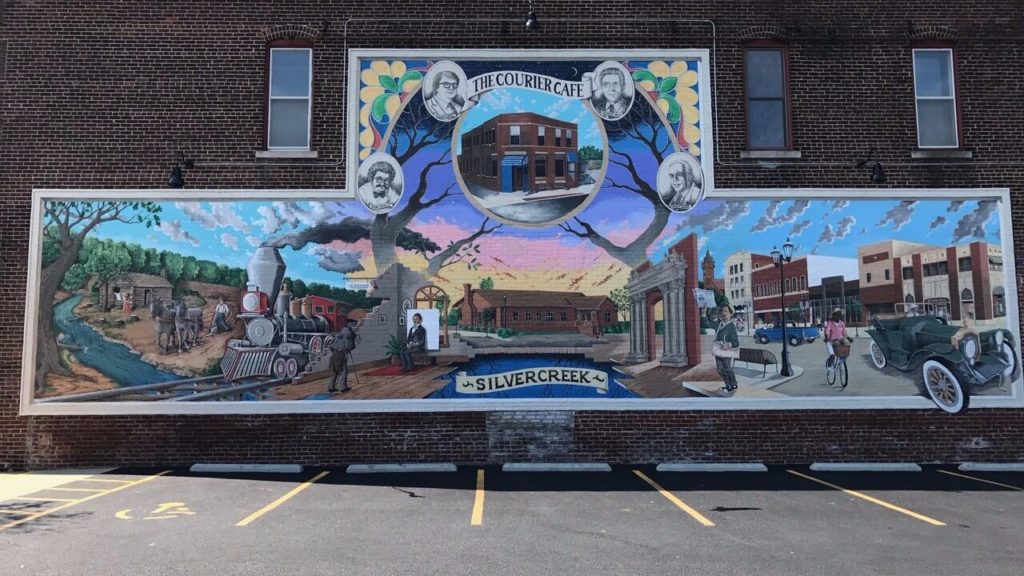 Mural on a brick wall. It shows three scenes: on the left a train moving through a pioneer settlement; center is a mill; on the right a downtown. The mural is colorful and represents different aspects of Urbana, Illinois.