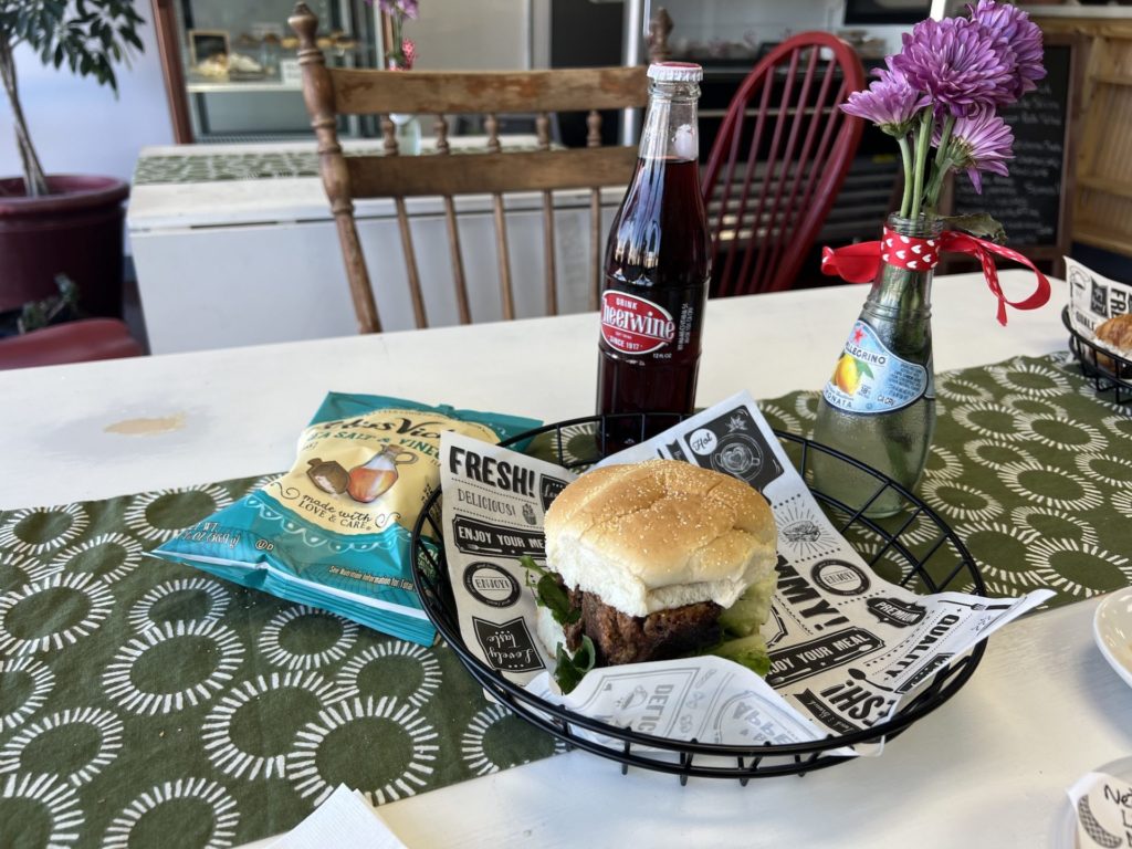 Meatloaf sandwich on a bun sits on a table with chips and bottled soda. 
