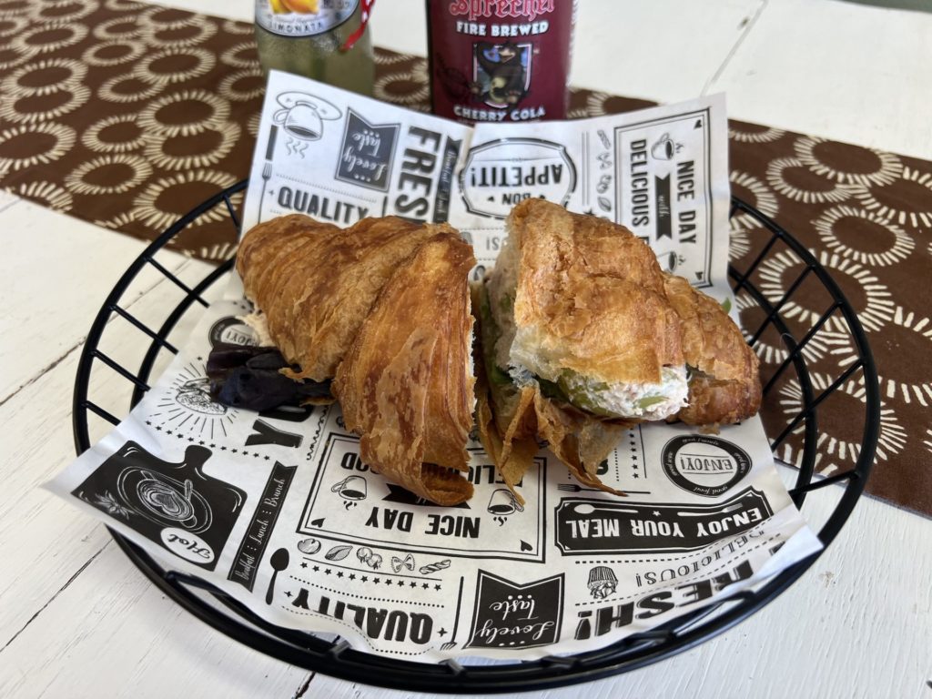 Chicken salad sandwich on a croissant sits in a wire basket with printed paper lining. 