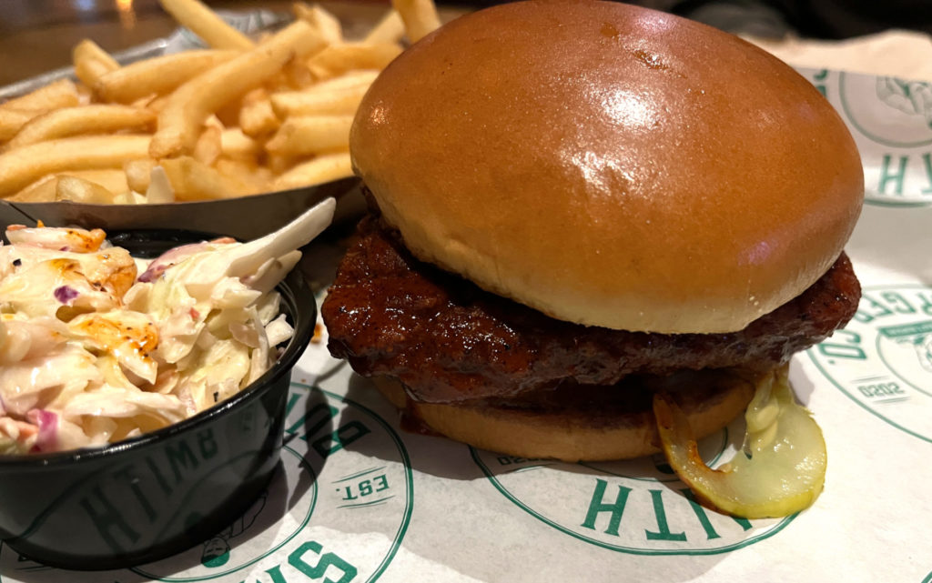 Fried chicken sandwich, dipped in Nashville hot sauce. On a platter is a side of mayo-based slaw and a side of French fries. There is a pickle sticking out of the bun on the sandwich.