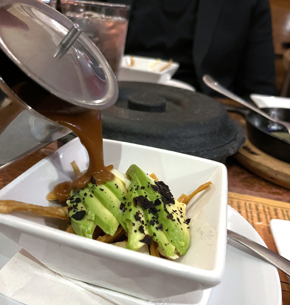 A white bowl with crispy tortilla strips and avocado slices pictured as pureed black bean soup is poured into the bowl from a metal container.
