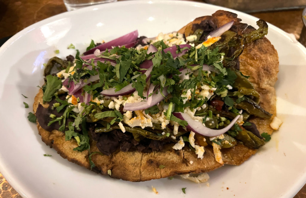 A large, plate-sized crispy and charred tortilla is topped with black beans, nopales (cactus), cheese, cilantro, red onion.