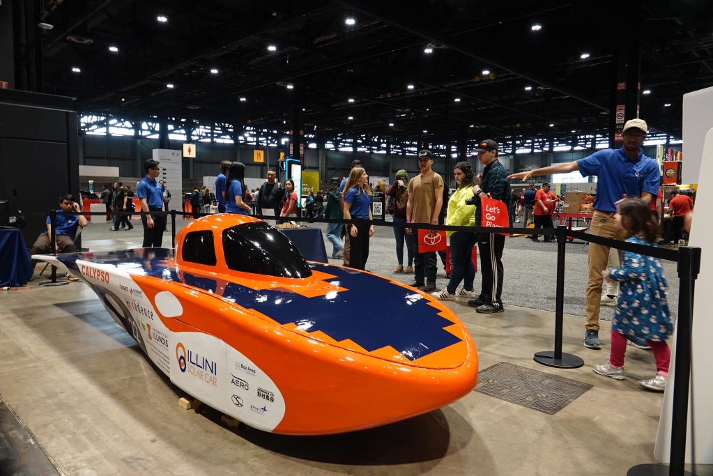 A blue, white, and orange boat shaped car sits on a cement floor in the Chicago Car Show. it is surrounded by a barrier with people staring at it on the other side.