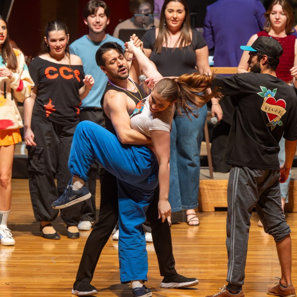 Performers wearing street clothes stand around a couple dancing. The man is wearing black jeans and a black tank top. The woman is wearing blue pants and a white top and has a long pony tail. Her left leg is lifted and she is twisting in the dance pose. Another performer stands to the right, wearing track pants, a black t-shirt, and a baseball cap.