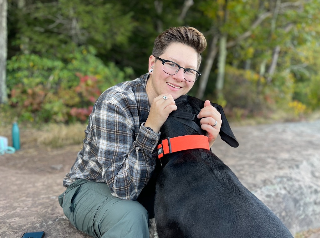 A woman with short brown hair and glasses sits outside near a forest. She its wearing a gray flannel shirt and jeans and scratching the ears of a black dog with an orange collar. 