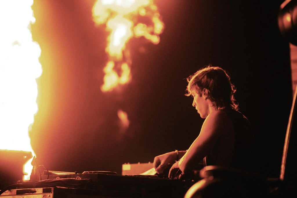 A side profile of a DJ intensely focused on the turntables during a performance, with dramatic flames erupting in the background, casting a warm, fiery glow over the scene. The image exudes the energy of a live electronic music concert.