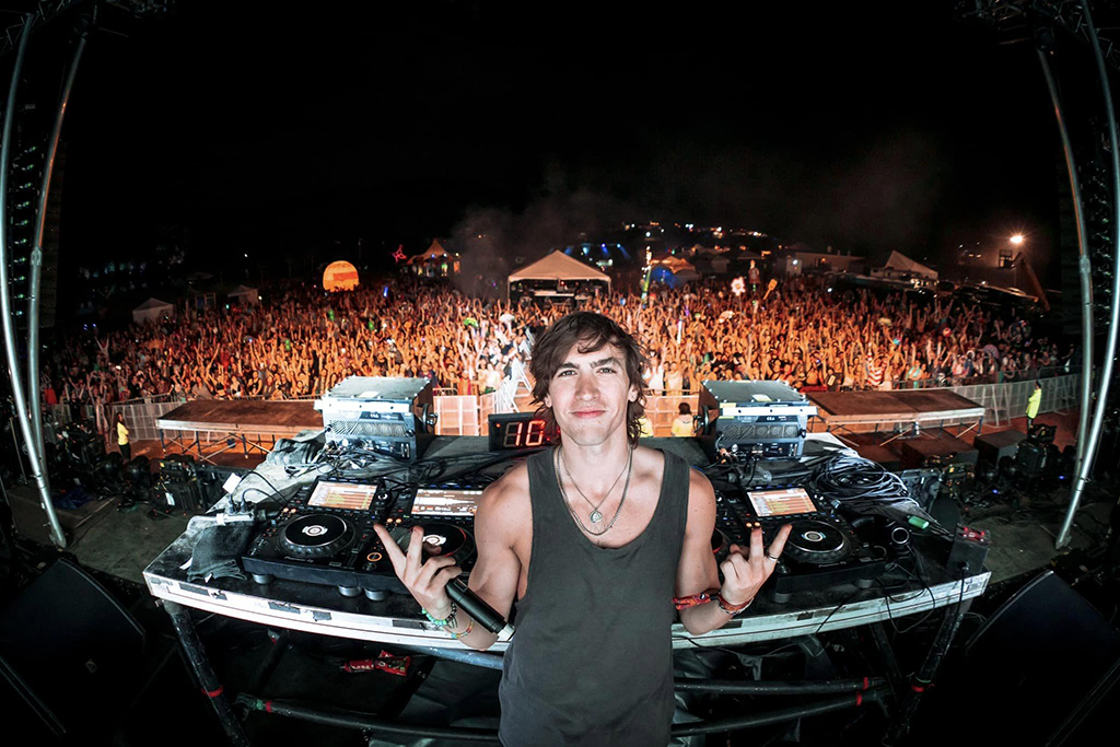 A DJ stands at a turntable setup, making a peace sign with their fingers, smiling at the camera with a crowd of festival-goers in the background. The image captures a moment of connection between the artist and the audience, set within a large outdoor music event.