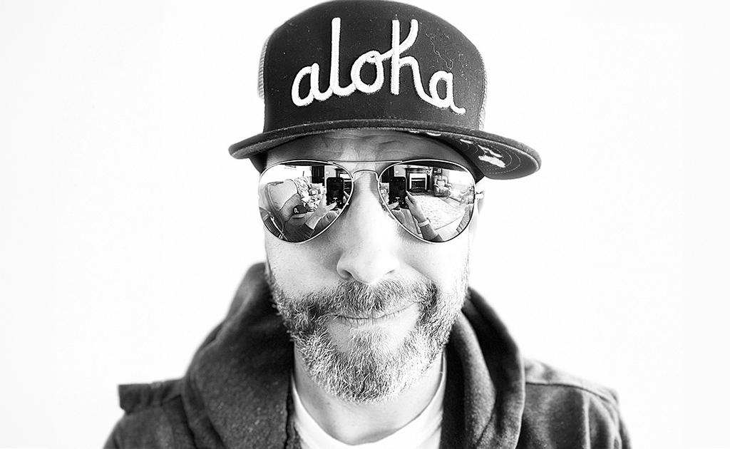 An individual is pictured wearing a cap with the word "aloha" on it, and reflective aviator sunglasses that show a mirrored scene. The photograph is taken in high contrast black and white, emphasizing the textural details of the cap and the person's facial hair. The individual's expression is relaxed and friendly.