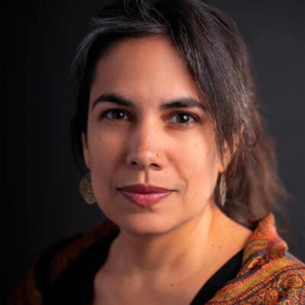 A Punjabi-Chicanx artist with brown skin, dark-brown eyes, and dark, gray-streaked hair pulled up into a loose twist looks into the camera. They are posed against a black background, wearing a black shirt, their grandmother’s orange patterned scarf, earrings, and a nose ring
