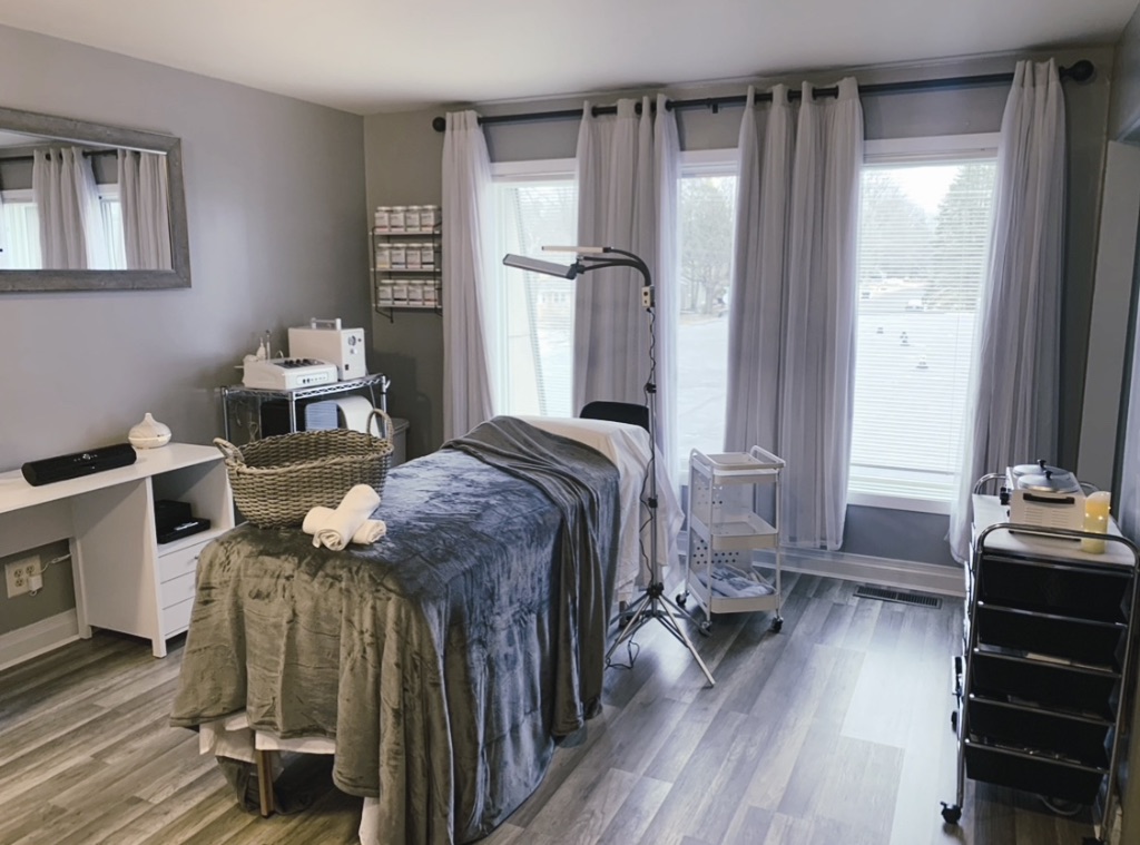 A light filled spa room with gray curtains and a bed with blankets. The room also has facial equipment, cabinets, and a grey plank floor.  
