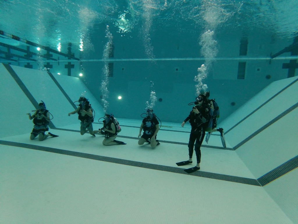 A group of scuba divers are all underwater in an indoor pool wearing full black diving gear. You can see black stripes on the bottom of the pool and bubbles floating up from their masks.