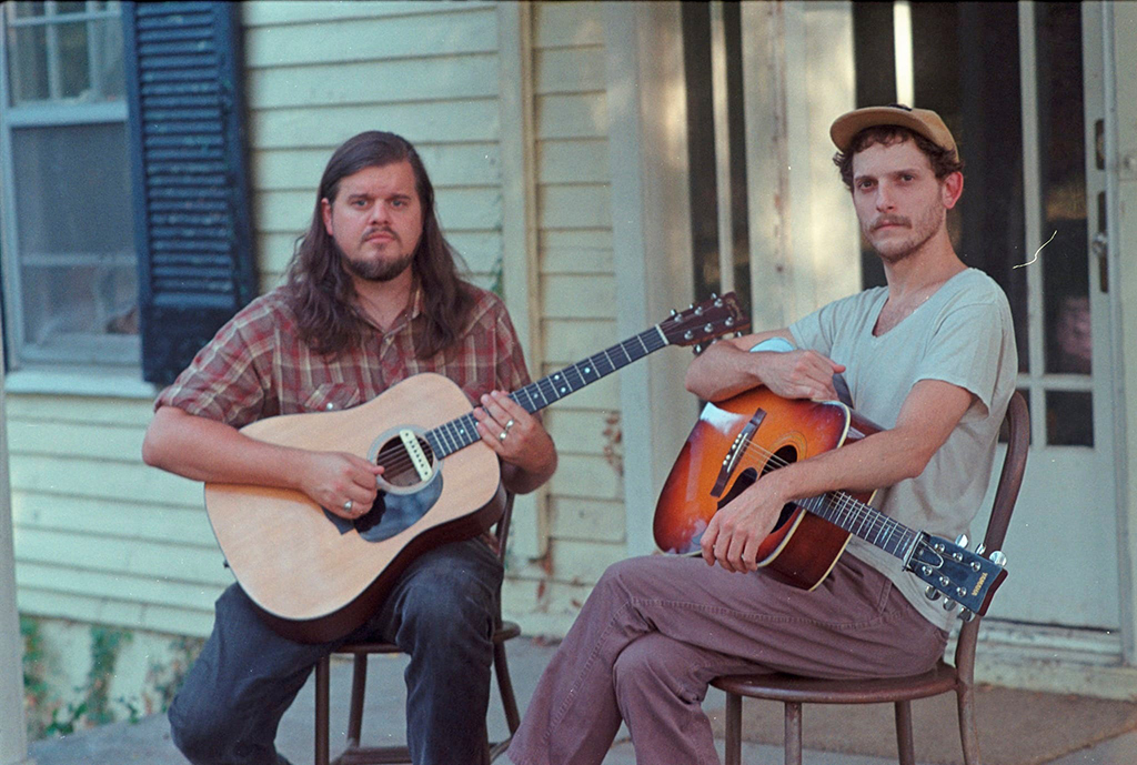 Two musicians are seated on a wooden porch, each holding a guitar, their expressions contemplative and serene. The setting is informal and homely, with one musician dressed in a plaid shirt and jeans, while the other dons a casual tee and cap. The rustic charm of the weathered house behind them, complete with traditional shutters and a white façade, frames this scene of camaraderie and creative partnership. The image conveys a sense of storytelling, a prelude to music that speaks of shared histories and authentic experiences.