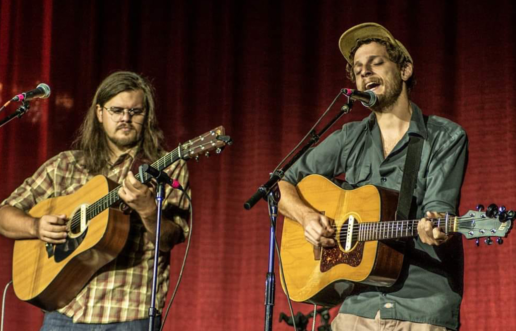 Two musicians stand side by side against a warm, red curtain backdrop, their expressions absorbed in the act of performance. The one on the left, with shoulder-length hair and a plaid shirt, gently cradles his guitar and focuses intently on his instrument. To the right, his bandmate, sporting a relaxed shirt and a cap, smiles as he sings into the microphone, his hands strumming his guitar with ease. The setting suggests an intimate concert, with each artist immersed in their craft, harmoniously delivering their music to an unseen audience.
