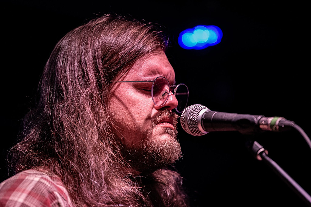 A close-up of a musician lost in the moment, his eyes behind round-framed glasses, almost closed as he pours his soul into the microphone. His long hair falls loosely around his shoulders, framing his face, and the plaid pattern of his shirt adds a touch of rustic authenticity to the scene. The warm stage lighting casts a passionate hue on the performance, with bokeh lights softly glowing in the background, emphasizing the intimate atmosphere of a live music setting.