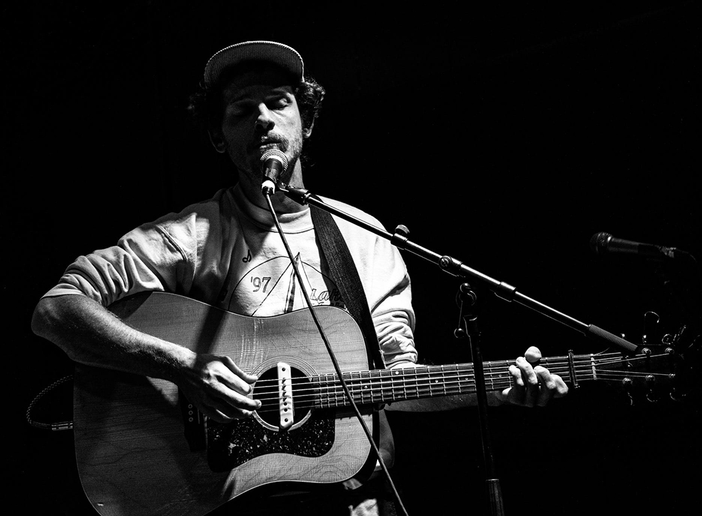 A musician is captured in a monochrome image, evoking a strong sense of focus and intensity. He stands alone, holding an acoustic guitar, fingers poised on the strings as if mid-strum. The musician is wearing a casual long-sleeved shirt, overlaid with a graphic tee, and a cap, suggesting a laid-back yet earnest persona. The microphone stands ready to catch the nuances of his performance, hinting at a scene filled with acoustic melodies. The absence of an audience in the frame turns all attention to the artist and his instrument, creating a private concert-like atmosphere.