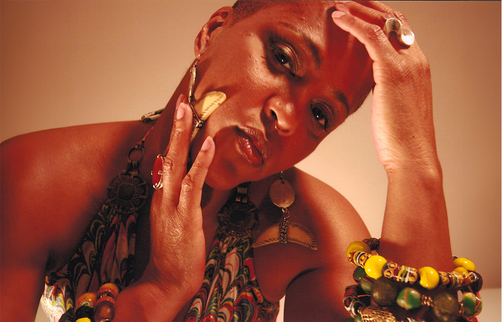 The individual in the photo is adorned with bold and colorful jewelry, including large earrings, multiple bracelets, and rings. They are striking a pose that suggests contemplation, with one hand placed gently on the side of their face. The warm lighting enhances the rich tones of their skin and the vibrant patterns of their attire, creating an intimate and artistic portrait.