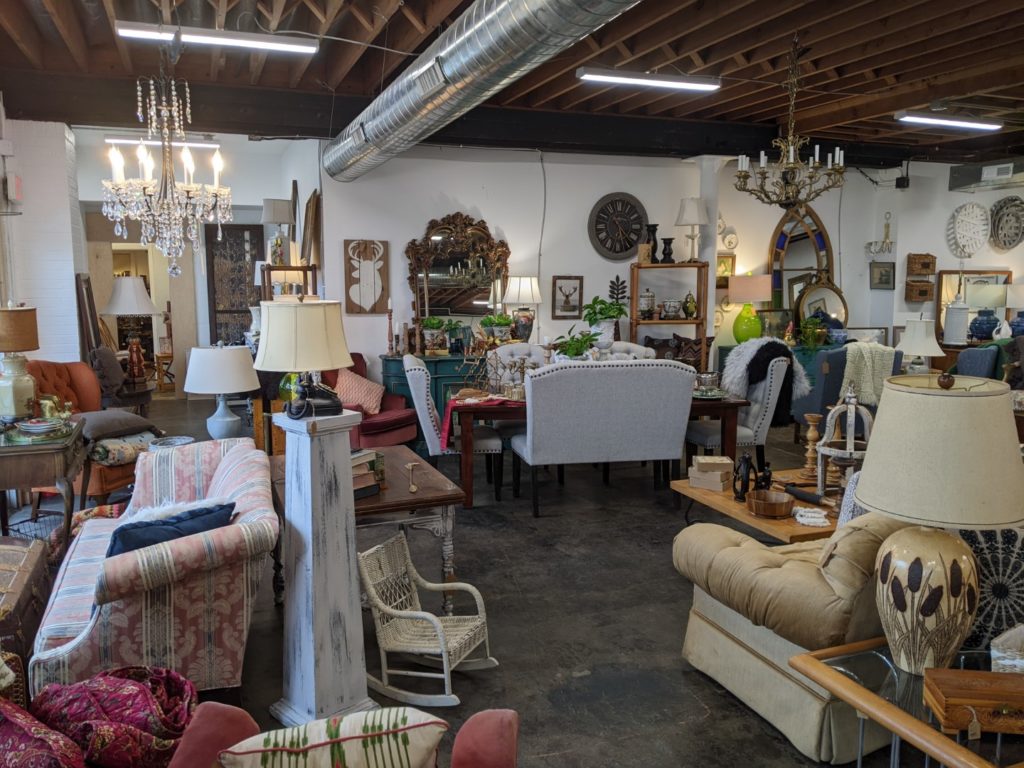 The inside of DIGS Consignment store, it is full of a variety of couches chairs, coffee tables, chandeliers, and lamps. And a dark cement floor is visible in the middle of the image.