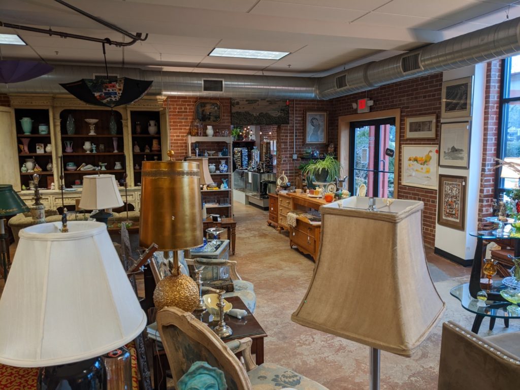 The inside of crossroads consignment store. it has low white ceilings, brick walls that are covered with framed pictures, shelves of vases, dressers, lamps, and an assortment of chairs and tables.