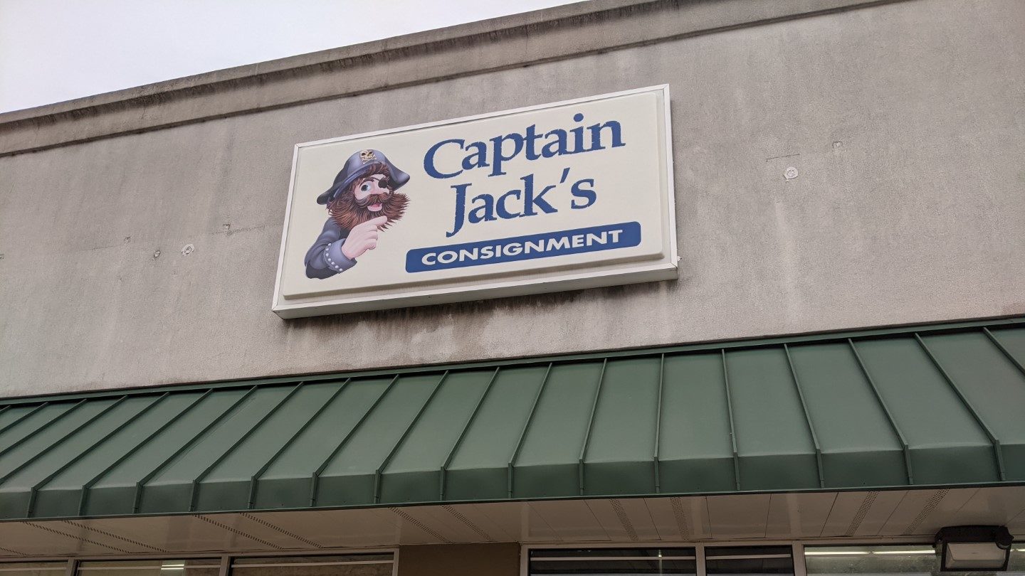 A concrete building with a green awning and a white sign with a pirate with a brown beard and blue jacket and hat and the words Captain Jack's consignment in blue letters. 