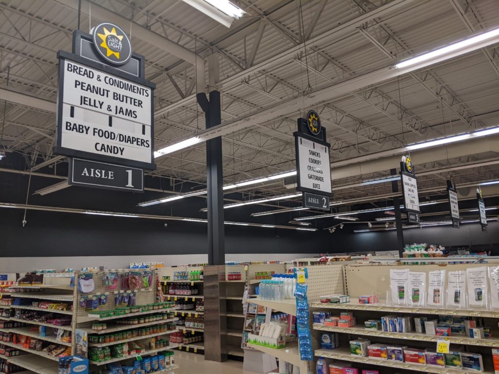The grocery section of the Salt and Light building. Shelves of groceries are visible with aisle signs hanging down from the ceilings. 