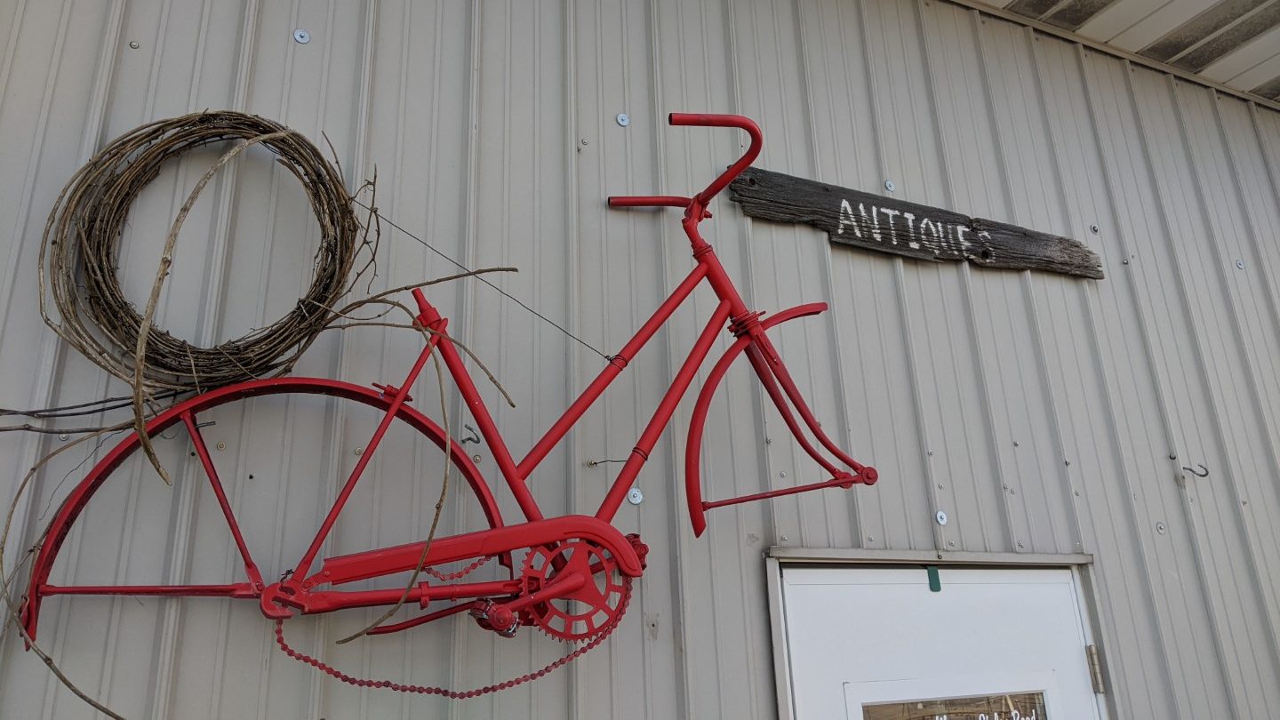 The side of a white metal building with half of an old red bicycle frame, a wreath, and a plank of wood with the word "antiques" painted in white over the white door.