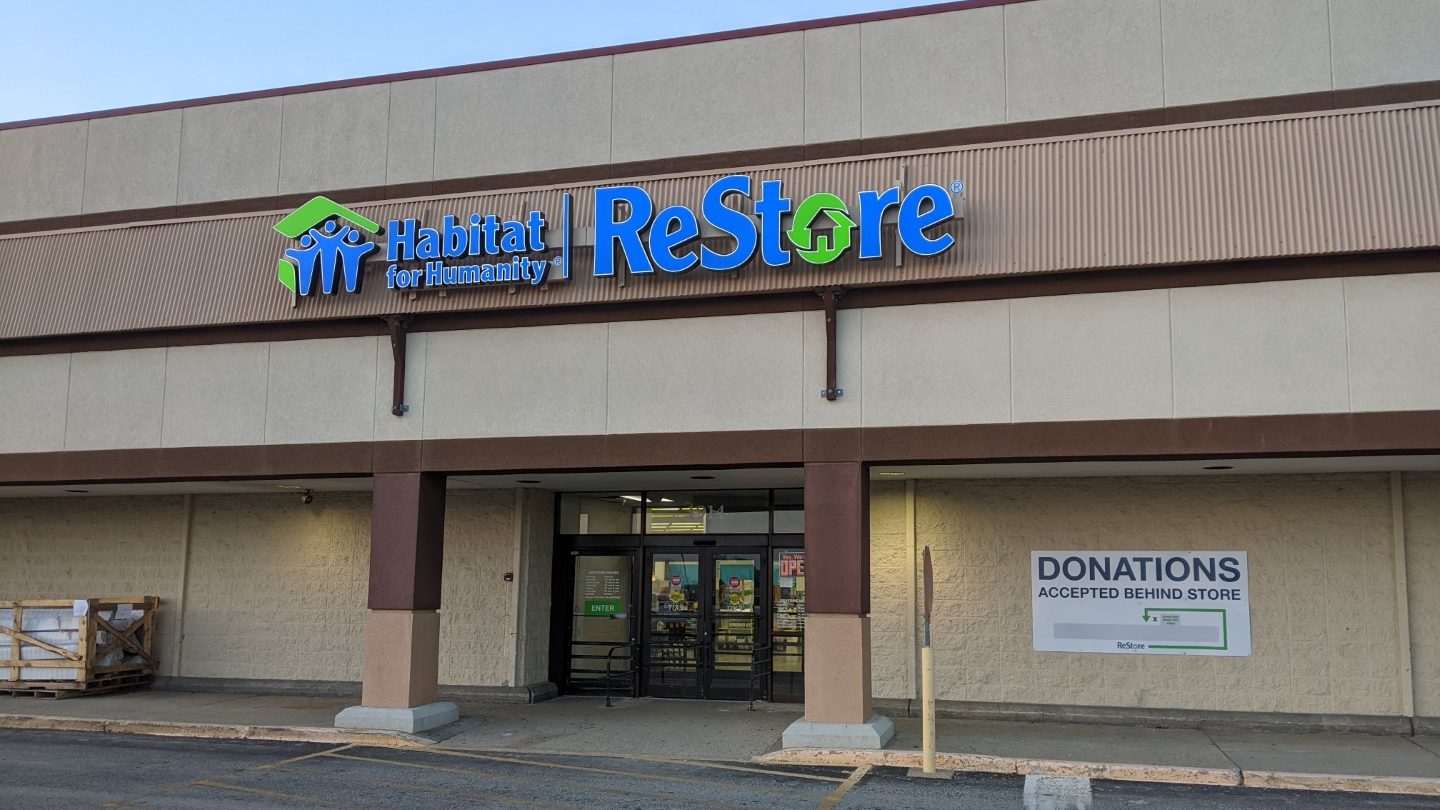 The outside of the Habitat for Humanity ReStore. The building is various shades of brown and tan with the letters in bright blue and green. 