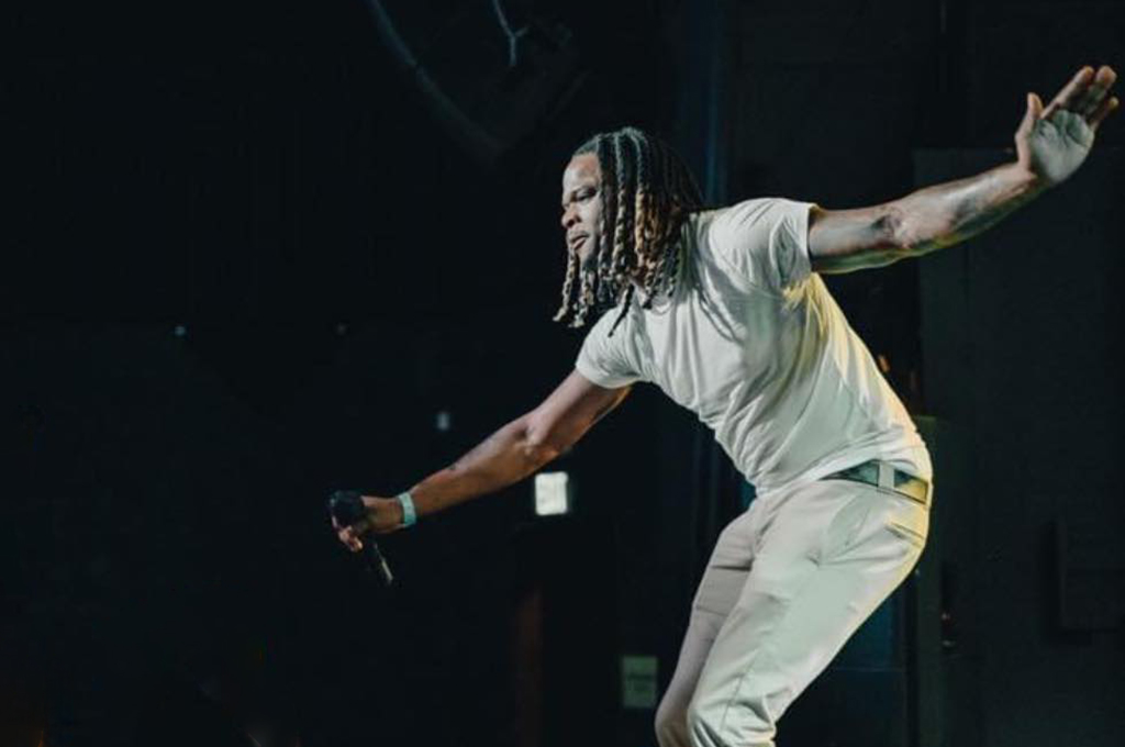 The image features a male performer in mid-motion on stage, dressed in a light-colored, fitted short-sleeve shirt and light-colored pants. His hair is styled in long dreadlocks that sway with his movement. He is holding a microphone in his left hand and has his right arm extended out to the side. The lighting casts a soft glow on him, highlighting his dynamic pose. The background is dark and nondescript, focusing the attention on the performer's actions.
