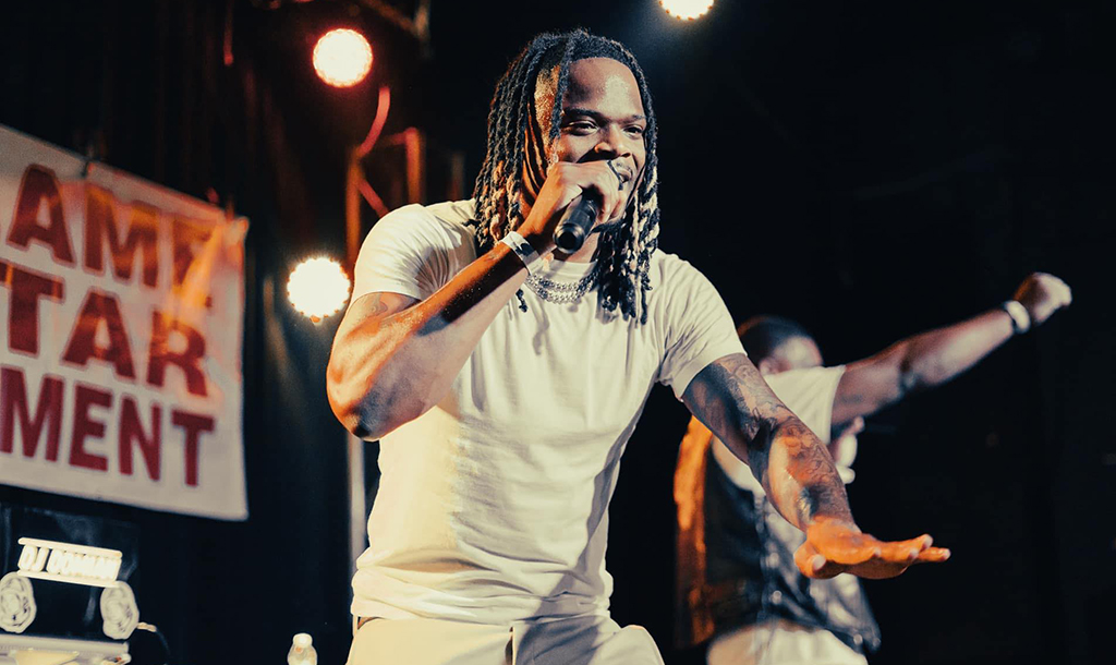 A person with long, dark dreadlocks is on stage performing. They are wearing a plain white t-shirt, which contrasts with the dark ink of tattoos visible on their arms. The person is holding a microphone in their right hand, gesturing openly towards the audience with their left hand. Behind them, a banner hangs with words in bold, red font. The backdrop is a dark curtain, and there are bright stage lights casting a glow on the scene, highlighting the performer and creating a dramatic ambiance.