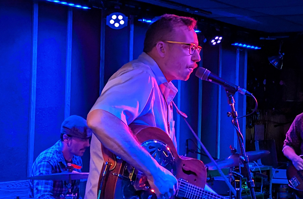 Two individuals are immersed in a musical performance on stage. The person in the foreground, clad in a light-colored shirt, is engrossed in playing an electric guitar. Behind them, another individual, donned in a checkered shirt and cap, is energetically playing the drums. The stage is bathed in a blue glow, casting an ambient light over the performers and their instruments. In the backdrop, curtains and stage lights add to the concert atmosphere.
