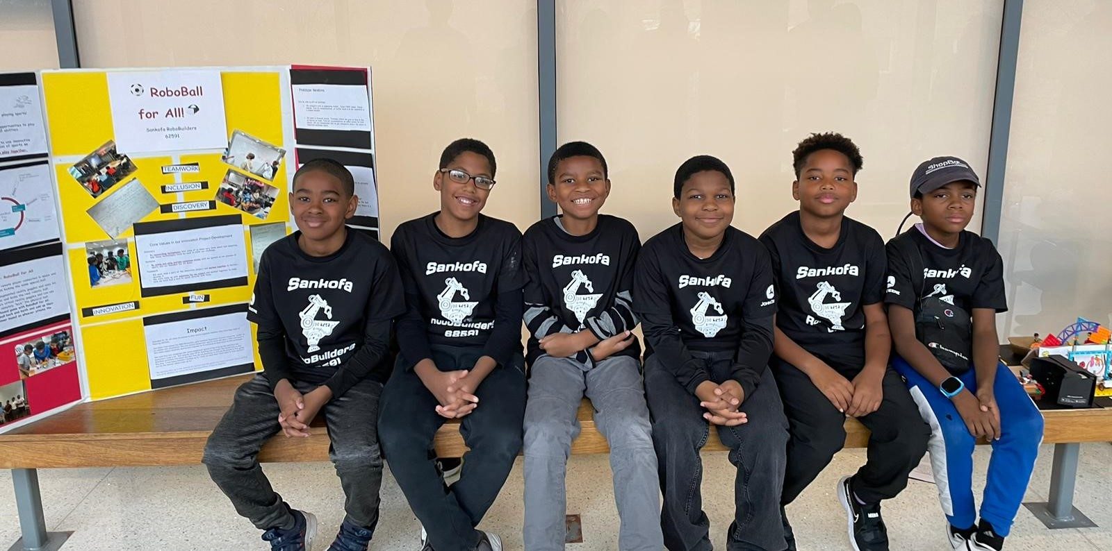 Six young Black boys are seating side by side on a bench. They are wearing black t-shirts with white print that say Sankofa RoboBuilders.