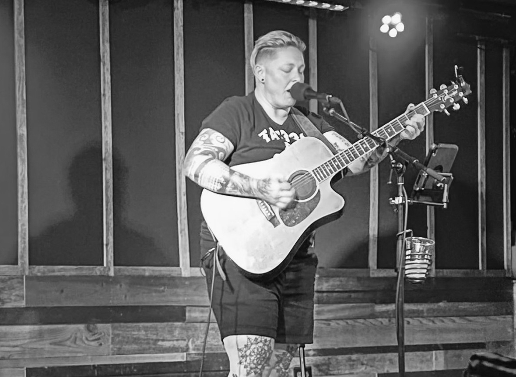 The black and white image features an individual performing on stage with an acoustic guitar. The musician has short-cropped hair and tattooed arms, which are visible as they wear a short-sleeved shirt. They are playing the guitar with focus and intensity, as evidenced by their closed eyes and the position of their mouth, suggesting they are singing into a microphone stand in front of them. The microphone and stand have an attached cup holder with a beverage cup in it. The performer is wearing dark shorts and a dark t-shirt with a visible graphic or text on the front. The stage has a rustic wooden backdrop, with vertical slats providing a textured background. The musician stands on what appears to be a wooden platform or stage, and the lighting creates soft shadows around them, emphasizing the intimate atmosphere of the live performance.