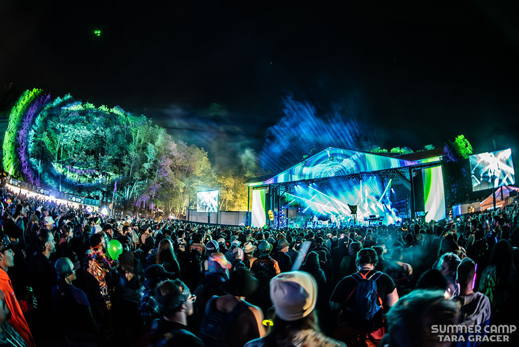 The scene captures a large gathering of people at an outdoor concert during the night. The crowd is dressed in various types of clothing, including hats and jackets, illuminated by the vibrant lights from the stage. The stage itself is covered and features bright lights in hues of blue and green, casting colorful reflections on the surrounding trees and attendees. Large screens on either side of the stage display live footage of the performance. The atmosphere is further enhanced by smoke or mist under the dark sky.