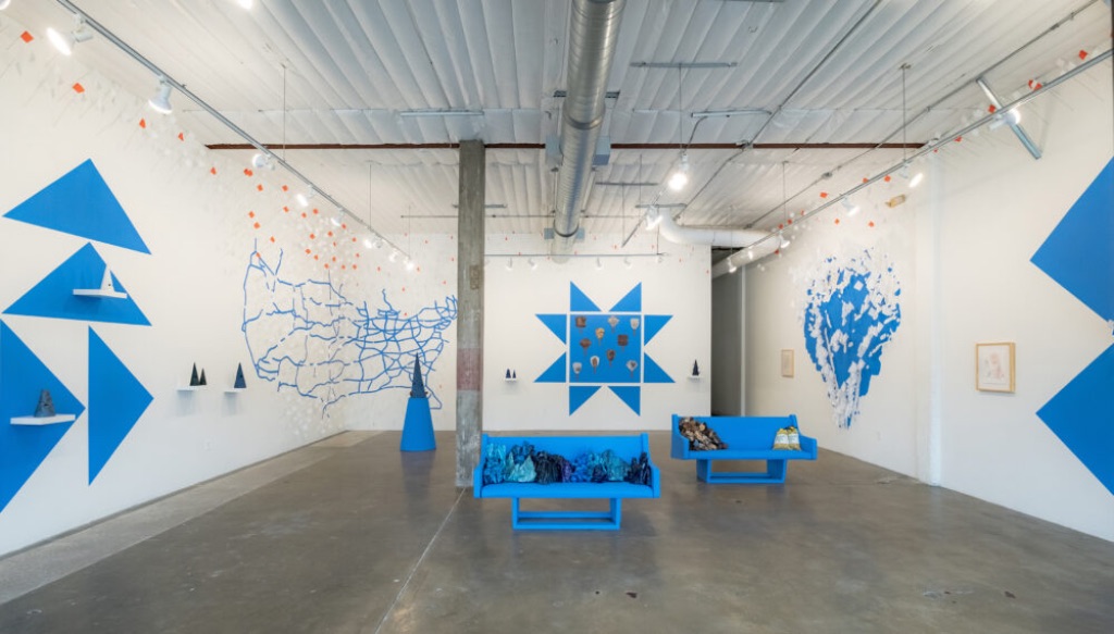 A gallery with white walls featuring a design of triangles, geometric shapes, an outline of the US, and an organic shape resembling an enormous puddle; all are painted in the same medium-blue color. Along the walls are shelves with cone-shaped sculptures and small orange-and-white flags. In the center of the space are two medium-blue benches covered with indecipherable objects