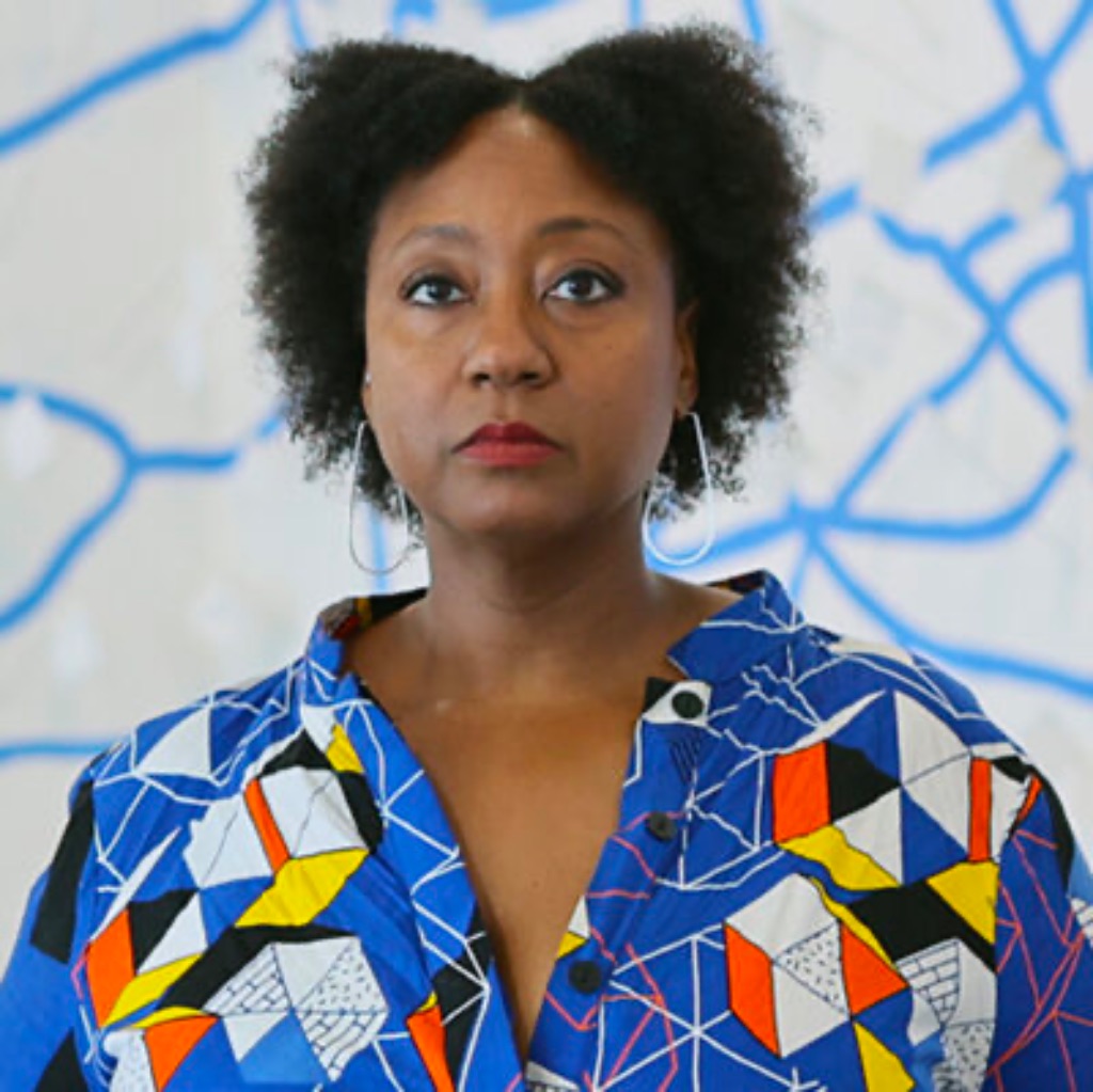 A Black woman with short hair looks out beyond the camera. She wears tear drop–shaped wire earrings, a colorful patterned shirt, and poses alongside a sculpture and mural