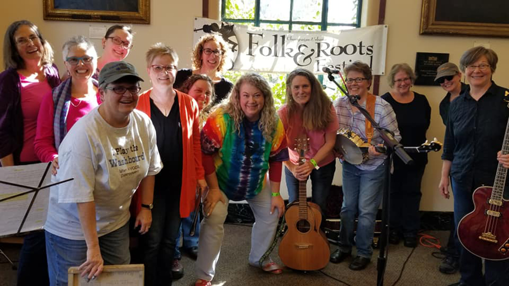 A group of individuals is gathered in a room with a banner reading "Folk & Roots Festival" in the background. They are dressed in casual attire, with several holding musical instruments such as a guitar and a banjo, suggesting they are a band or a group of musicians. The setting has a communal and informal feel, with visible music stands and microphones, indicating a performance or a rehearsal space.