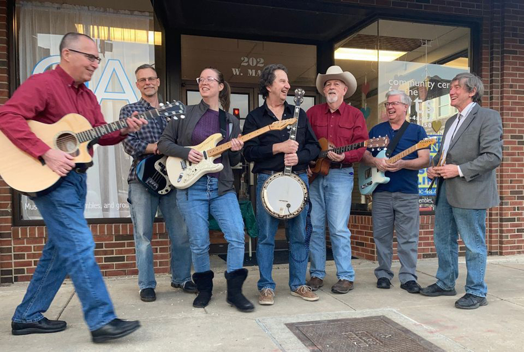 A group of seven individuals stands in front of a brick building with large windows and a sign that reads "Community Center for the Arts." They are casually dressed, with some wearing jeans and others in slacks, paired with a variety of tops from button-up shirts to a pullover. Each person is holding a musical instrument: there are two acoustic guitars, two electric guitars, a banjo, and a bass guitar. The group appears in a candid moment, some members smiling and engaging with each other, creating a sense of camaraderie and joy. The setting suggests they may be a band preparing for or having just completed a performance.