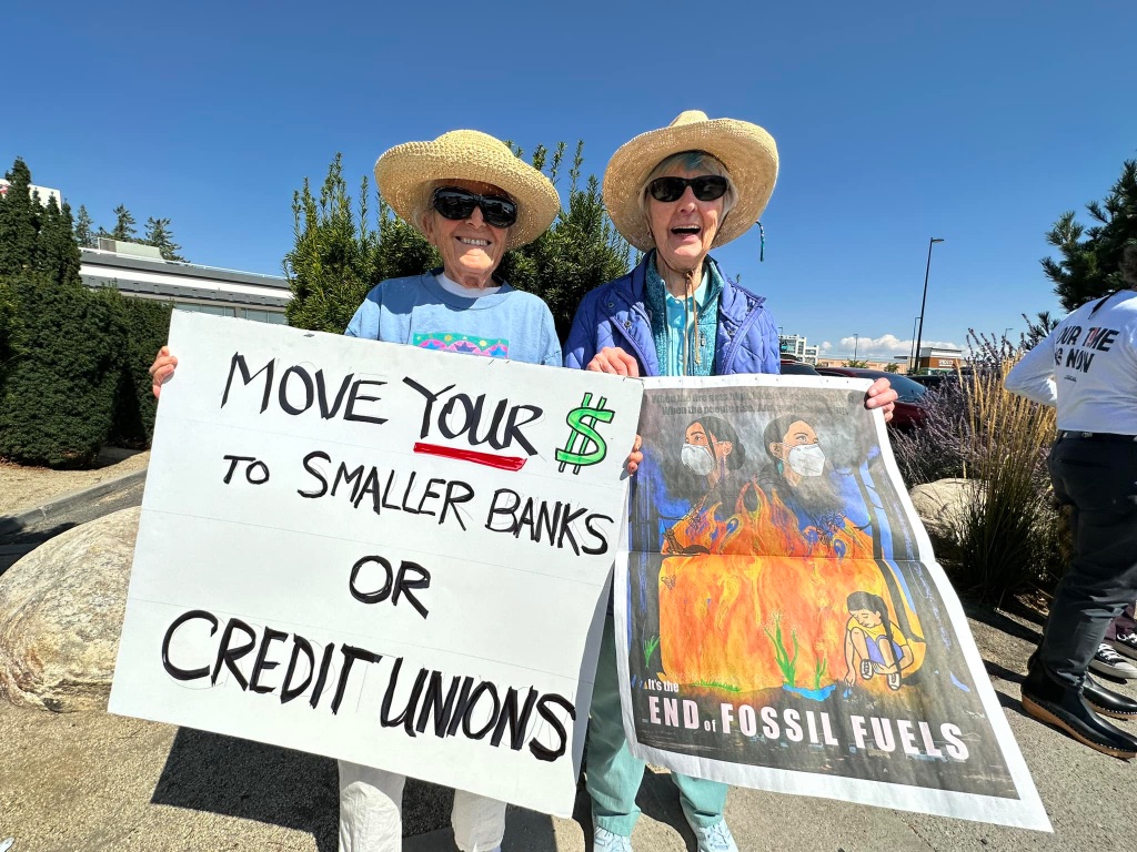 Two older white women are wearing big straw hats, sunglasses and standing on a sidewalk holding protest signs.