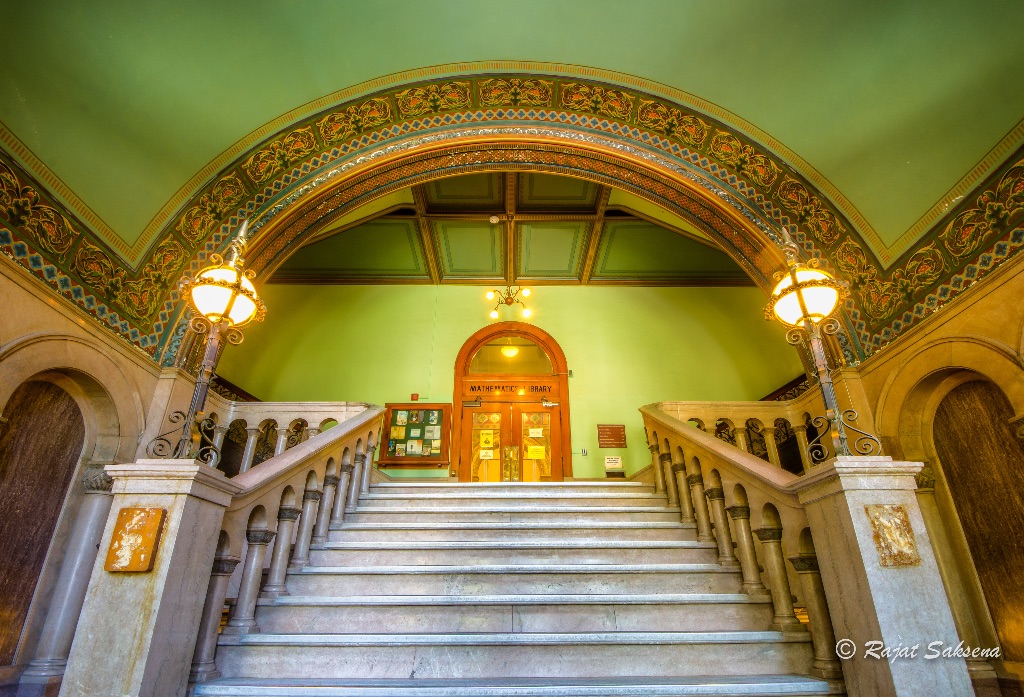 A picture of the staircase of altgeld hall. The ceiling is green with intricate painted ceiling and a wooden door with an arched doorway. 