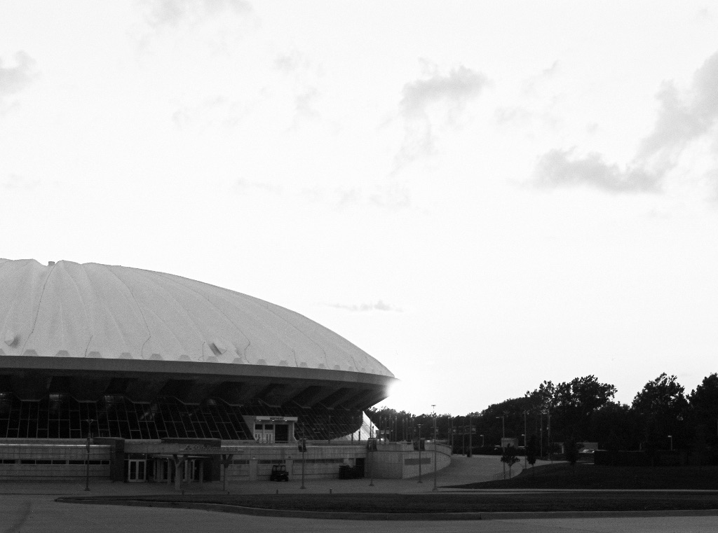 A black and white picture of the State Farm center. It sits on the left half of the picture with the sun shining behind it. There are small clouds in the distance. 