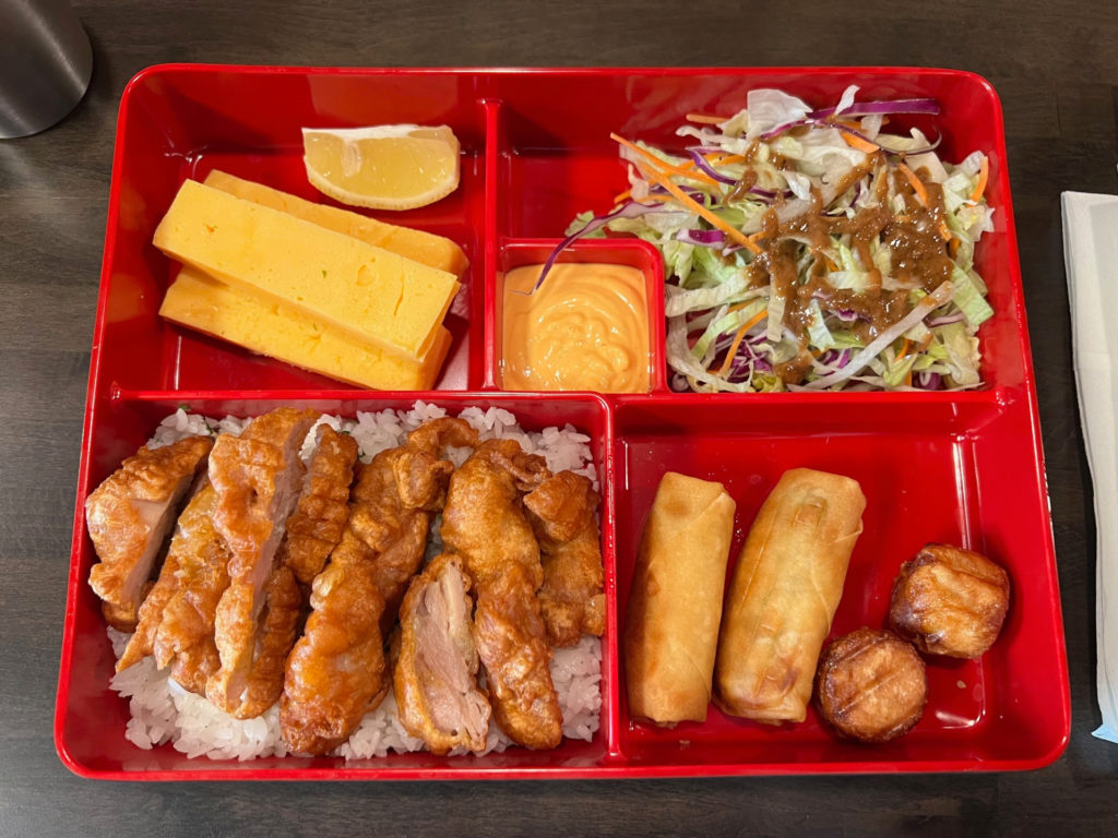 A red bento box of chicken katsu with tamago, salad, egg rolls, and fish cakes.